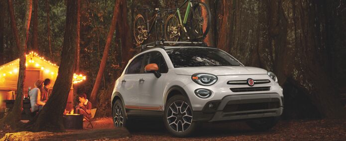 A 2021 Fiat 500 loaded with two bikes on an available roof rack, parked in the woods beside a cabin.
